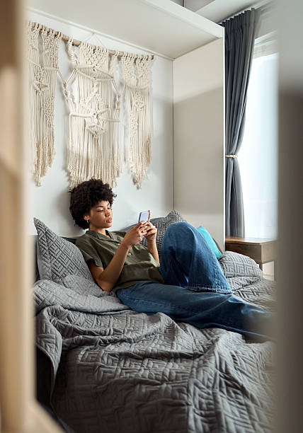 istock Young African American woman using phone at home sitting on bed, vertical. 2214671473