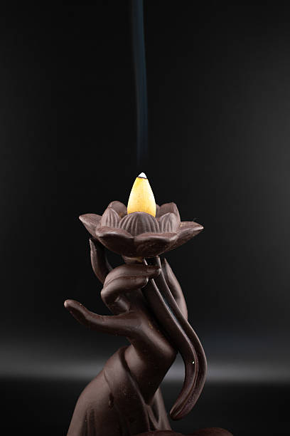 istock Tranquil scene of a dark ceramic backflow incense burner shaped like a hand holding a lotus, with a lit incense cone releasing delicate smoke. Evokes peace and aromatherapy 2214674458