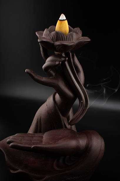istock Tranquil scene of a dark ceramic backflow incense burner shaped like a hand holding a lotus, with a lit incense cone releasing delicate smoke. Evokes peace and aromatherapy 2214674486