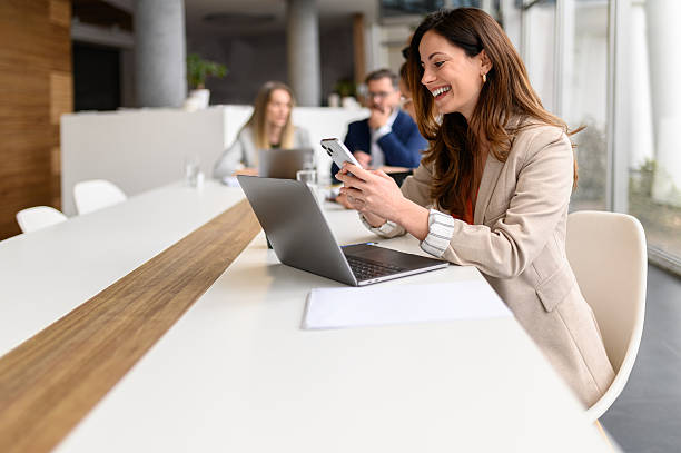 istock Smiling female lawyer texting over mobile phone while working over laptop in board room with colleagues in the background 2214679644