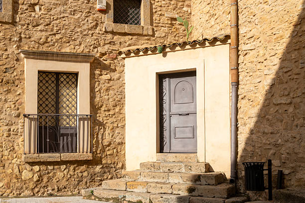 Niscemi, Sizilien - Italy - 03-26-2025: Sunlit stone facade in Niscemi featuring an iron door with steps, metal balcony, barred windows and a cactus plant on a tiled roof corner.