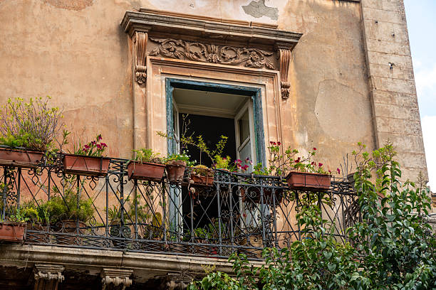 Ragusa, Sizilien - Italy - 04-03-2025: Ornate stone-framed window with open shutters and wrought iron balcony decorated with flower pots, located on a weathered facade in Ragusa.