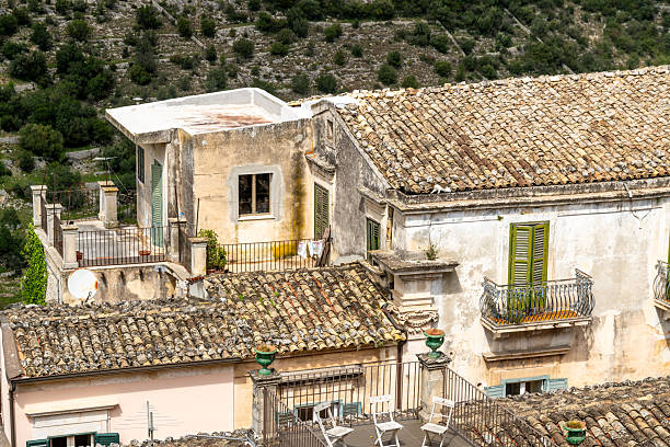 Ragusa, Sizilien - Italy - 04-03-2025: Aged tiled rooftops and weathered balconies with green shutters overlook a hillside landscape in the historic town of Ragusa, Sicily, under sunny skies.