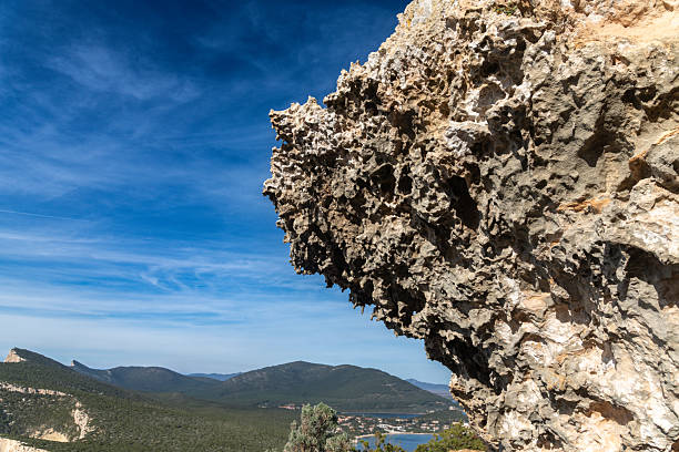 Eroded limestone cliff with rugged surface textures rising above a green valley, under a vivid blue sky with distant mountain landscape.