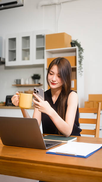 istock Happy Asian woman packing parcel boxes at a sofa table, smiling joyfully while checking online orders on laptop.She prepares products for delivery,managing her successful home-based online business 2214704318