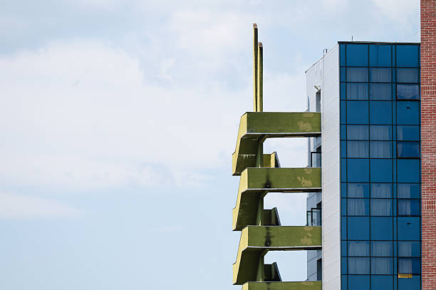 A partial view of a contemporary building showcasing bold green external staircases protruding from the side, alongside sleek blue glass windows and red brick. The mix of materials and colors creates a striking architectural contrast, with the stair structure adding a sculptural, geometric element to the composition.
