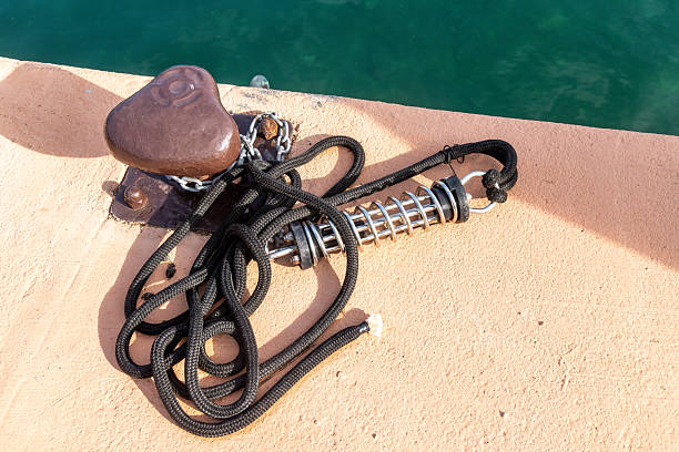 A metal dock cleat secured with a black rope, a steel mooring spring, and a chain is fixed on a pier above turquoise water.