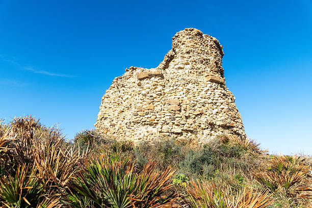Old stone watchtower on a coastal cliff in SArchittu, Sardinia, surrounded by rugged rocks, low vegetation, and a clear blue sky.