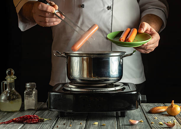 istock A chef is skillfully dropping sausages into a pot on the stove while holding a green plate with more sausages. Ingredients are scattered on the kitchen counter 2214708046