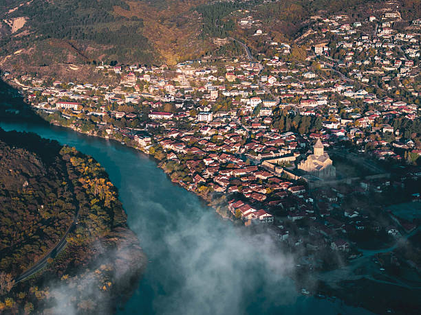 A stunning aerial view captures the historic town of Mtskheta, Georgia, situated at the confluence of the Aragvi and Kura rivers. The ancient Svetitskhoveli Cathedral stands prominently amidst the town's traditional red-roofed houses. Mist rises from the meeting of the turquoise rivers, creating an ethereal atmosphere. The surrounding landscape features rolling hills and lush greenery, highlighting the historical and natural significance of this UNESCO World Heritage site.