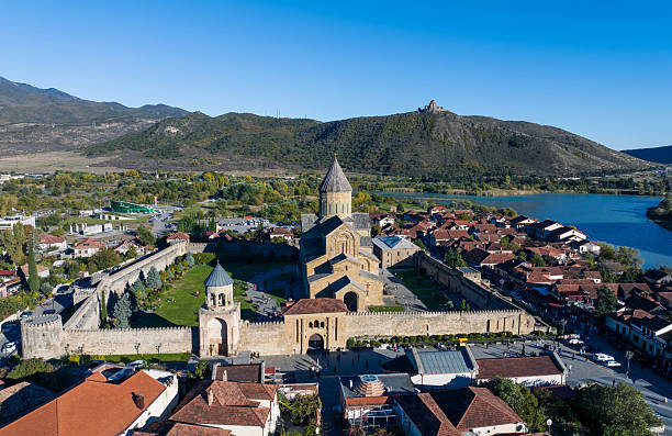 A stunning high-angle aerial photograph captures the magnificent Svetitskhoveli Cathedral in Mtskheta, Georgia, surrounded by its defensive walls and nestled within the town's traditional red-roofed houses and lush greenery. The cathedral's impressive architecture and historical significance are evident in this detailed view. Roads and trees meander through the town, highlighting the importance of this UNESCO World Heritage site in Georgian history and culture.