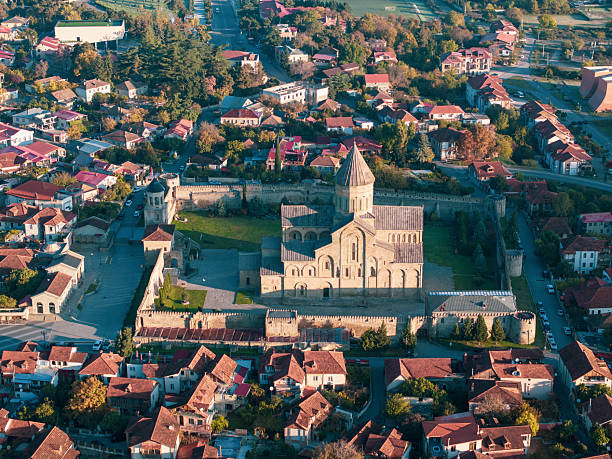 A stunning high-angle aerial photograph captures the magnificent Svetitskhoveli Cathedral in Mtskheta, Georgia, surrounded by its defensive walls and nestled within the town's traditional red-roofed houses and lush greenery. The cathedral's impressive architecture and historical significance are evident in this detailed view. Roads and trees meander through the town, highlighting the importance of this UNESCO World Heritage site in Georgian history and culture.