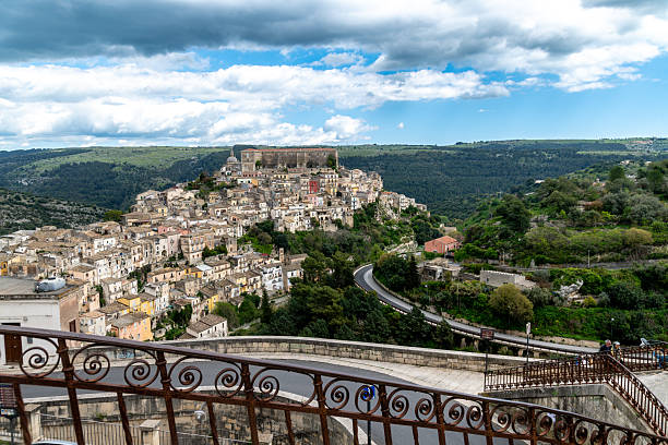 Historic Ragusa Ibla with stone houses and winding roads, surrounded by green hills and a partly cloudy sky, seen from a hilltop viewpoint.