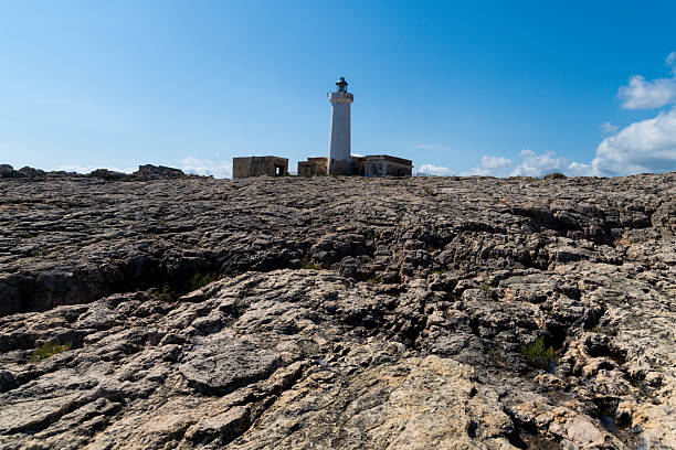 Lighthouse near Syracuse standing on a wide, rugged rock surface under blue sky, with small abandoned stone buildings beside the main struct