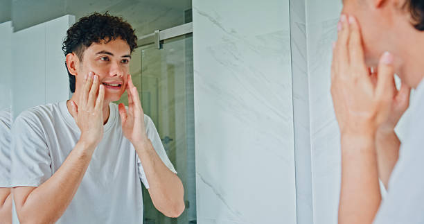 istock Young man smiling while applying moisturizer to his face in front of mirror in the bathroom at home. Healthy lifestyle in morning routine concept. 2214720089