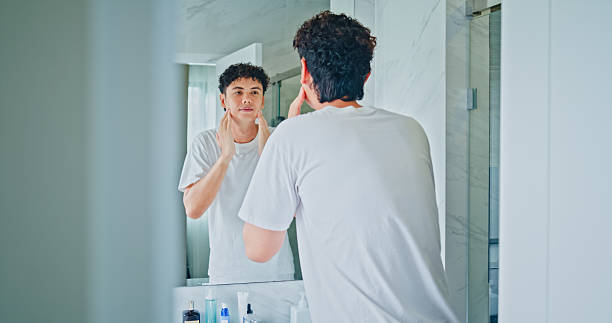 istock Young man smiling while applying moisturizer to his face in front of mirror in the bathroom at home. Healthy lifestyle in morning routine concept. 2214720093
