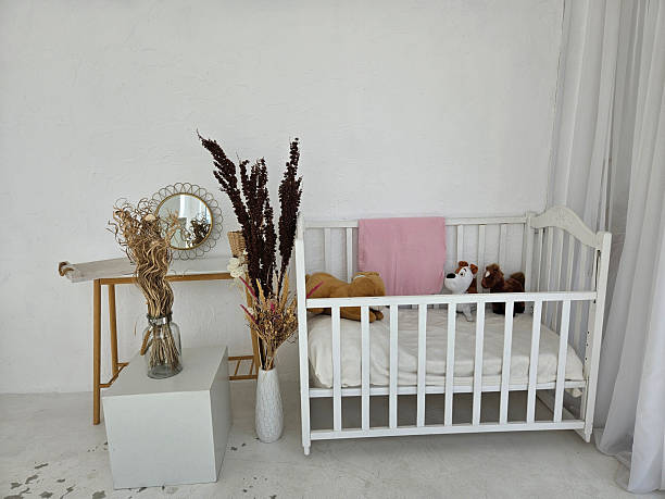 Baby cot with soft toys against white wall with changing table and mirror, also vases with dried flowers