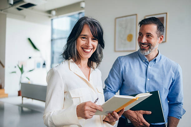 A confident woman holding a notebook and pen, accompanied by a smiling professional male colleague, working together in a bright and modern office environment.