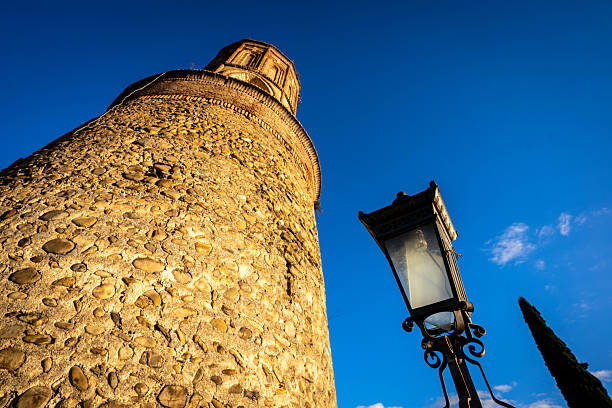 A charming street scene in Sighnaghi, Georgia, captured in warm sunlight, features a brick building with a window reflecting the sky in the foreground. A traditional stone bell tower rises prominently in the background against a clear blue sky with a few wispy clouds. A narrow road winds past wooden fences and greenery, showcasing the historic architecture and atmosphere of this town in the Kakheti region