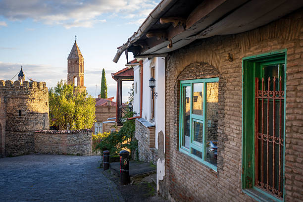 A picturesque view captures a cobblestone street winding through Sighnaghi, Georgia, with a mix of brick and stone buildings lining the way. A prominent stone bell tower rises in the background against a partly cloudy sky. Colorful window frames and traditional architectural details add to the charm of this historic town in the Kakheti region, likely taken in the soft light of early morning or late afternoon.