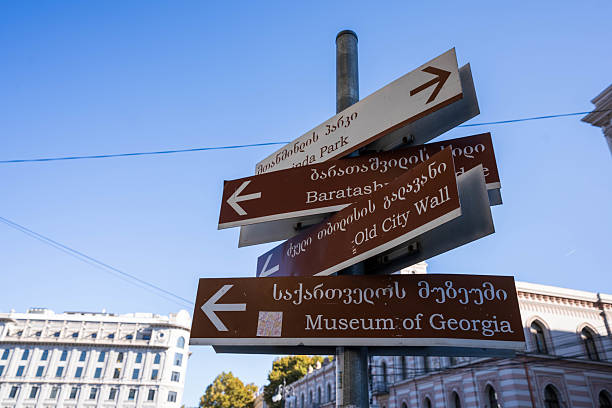 A vibrant photograph captures a multi-directional signpost in Tbilisi, Georgia, displaying names of various locations and landmarks in both Georgian and English. The signpost stands amidst lush greenery under a clear blue sky, providing guidance for locals and tourists navigating the city.