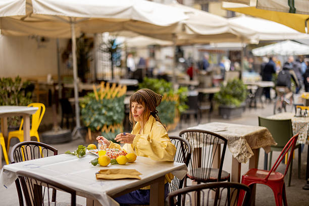 A woman in a yellow jacket enjoys a plate of pasta alone at a street cafe under large umbrellas. This image reflects the charm of solo travel, slow food culture, and authentic local dining