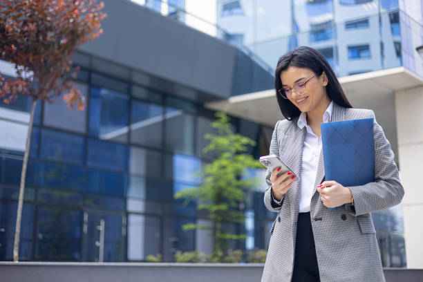 istock Smiling businesswoman holding a folder while using a smartphone, standing in front of a modern office building in a bustling city 2214739117
