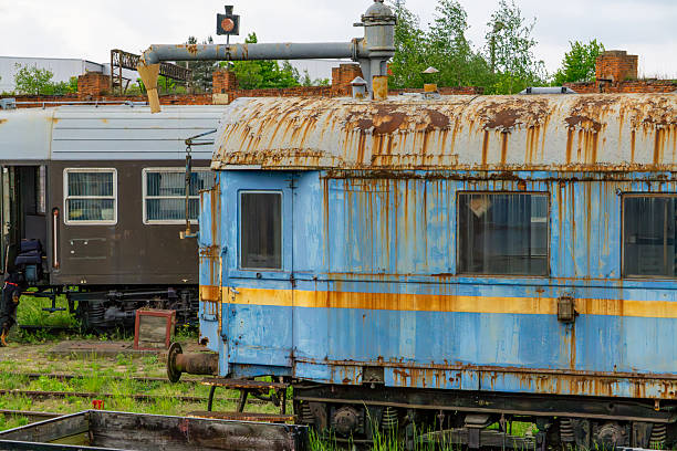 Old, rusty blue railway car with a yellow stripe, standing on a railway siding. Visible signs of corrosion and wear suggest long-term disuse. In the background, a newer train car and elements of railway infrastructure can be seen. The image captures the atmosphere of decaying technology and abandoned rail equipment.