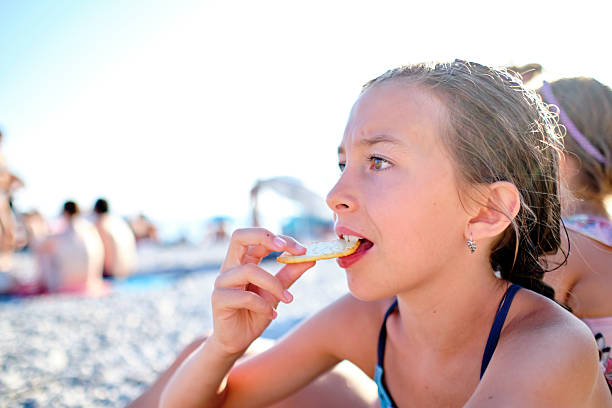 A young girl enjoys a quiet, sun-drenched moment while eating a crisp snack on a pebble-covered beach, her expression reflecting the simple joys of summer. The soft light and relaxed setting convey warmth, innocence, and the carefree essence of childhood holidays.