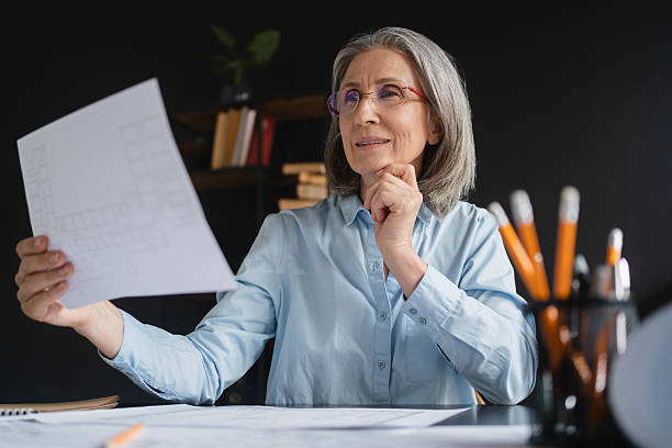 istock Caucasian mature woman architect sitting at her office desk with a desktop computer, working with construction plans, holding a sheet of paper, and looking at a creative design project 2214753636