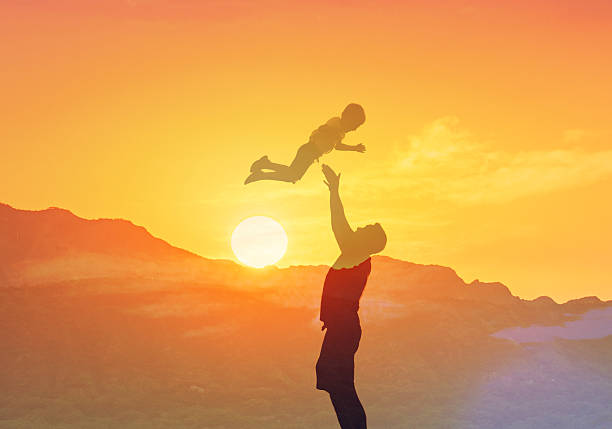 A father joyfully lifts his child in the air as the sun sets on a tranquil beach, capturing a moment of playful bonding and happiness, with water glistening around them.