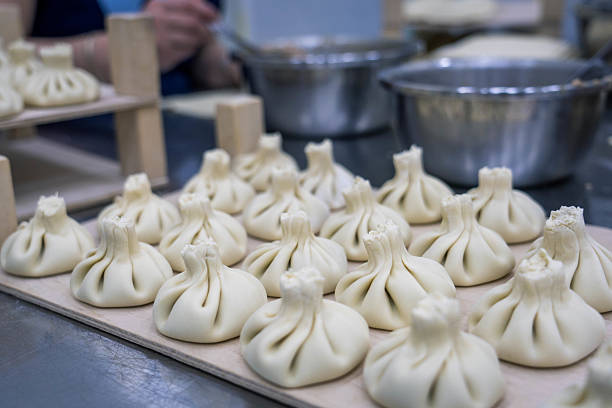 A close-up shot captures rows of uncooked khinkali, traditional Georgian dumplings, neatly arranged on a wooden surface, likely in a restaurant or kitchen in Tbilisi, Georgia. The intricate pleats at the top of each dumpling are clearly visible. In the blurred background, stainless steel bowls and hands suggest the ongoing process of preparing this popular Georgian dish