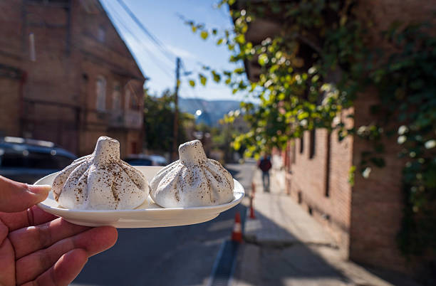 A close-up shot captures rows of uncooked khinkali, traditional Georgian dumplings, neatly arranged on a wooden surface, likely in a restaurant or kitchen in Tbilisi, Georgia. The intricate pleats at the top of each dumpling are clearly visible. In the blurred background, stainless steel bowls and hands suggest the ongoing process of preparing this popular Georgian dish.