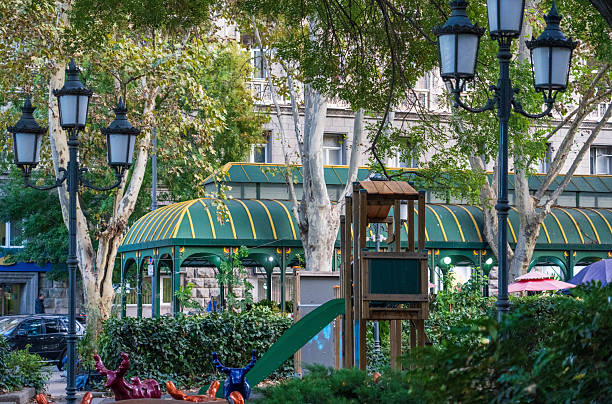 A charming scene within a Tbilisi park, Georgia, features a weathered wooden park bench in the foreground, with whimsical, colorful sculptures of reclining figures nearby. Beyond the bench, a playground structure with a green slide is partially visible, surrounded by lush greenery and trees. Elegant, vintage-style street lamps stand nearby, and a classic building rises in the background under a soft, overcast sky, suggesting a peaceful urban oasis
