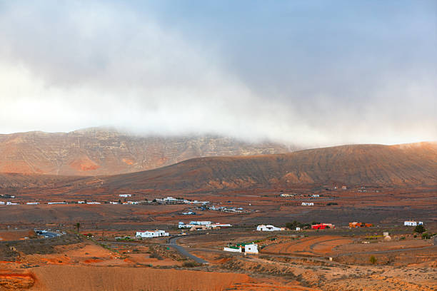 Typical Canary Islands landscape with rugged brown mountains shrouded in low clouds, creating dramatic and misty atmosphere. Scattered white houses dot the arid terrain of picturesque rural scenery