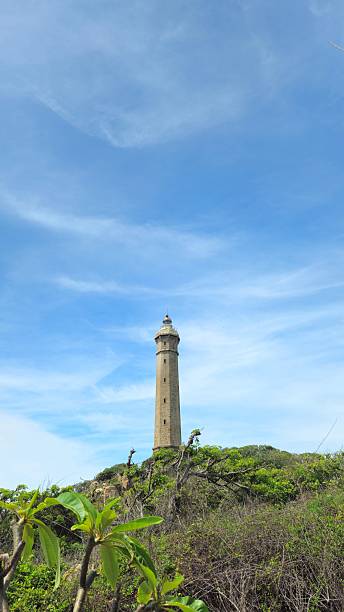 Ke Ga Lighthouse or Khe Ga Lighthouse rises above lush green trees under a bright blue sky in Binh Thuan province, Vietnam.