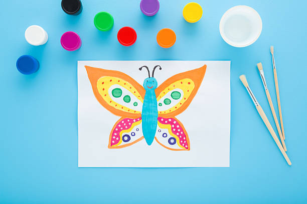 istock Painted colorful butterfly on white paper with gouache colors on light blue table background. Pastel color. Closeup. Top down view. 2214811205
