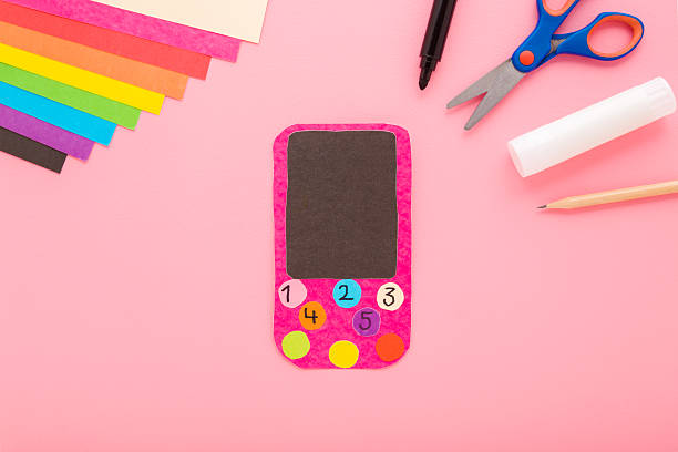 istock Smartphone, glue, scissors, marker, pencil and colorful application paper on light pink table background. Pastel color. Creating child gadget for playing. Closeup. Top down view. 2214811489