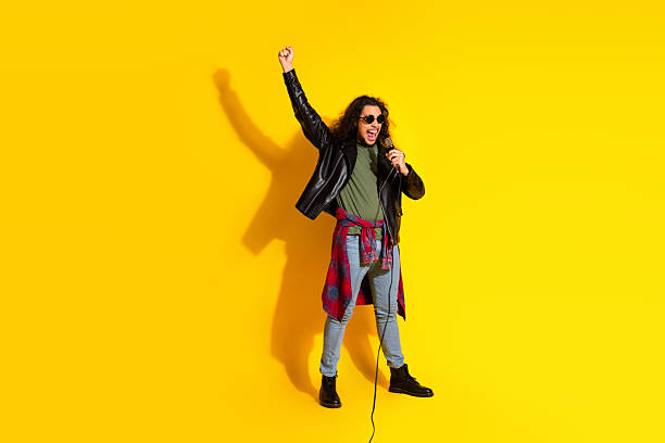 istock Young man in leather jacket performing with microphone against vibrant yellow background, expressing energy and style 2214827779