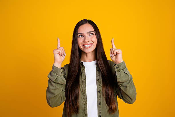 istock Happy young woman pointing up with both hands while smiling, posing in casual attire against a vibrant yellow background 2214828559