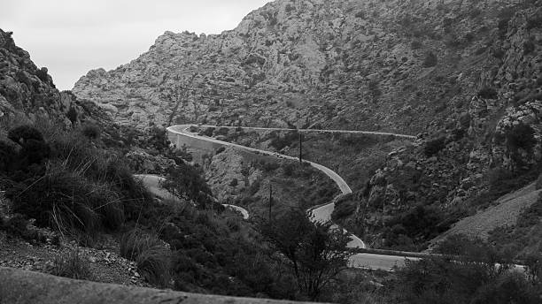 Black and white photo of a winding road cutting through rugged mountain terrain in Mallorca, Spain. A tranquil and timeless landscape with artistic contrasts and natural beauty.