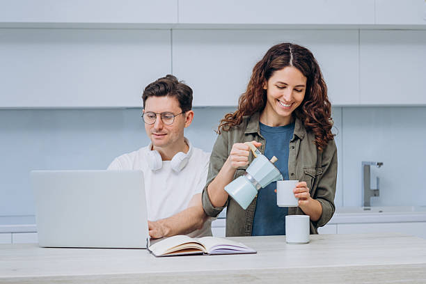 istock Woman pouring coffee while man works on laptop, both smiling in bright, modern kitchen setting. 2214830679