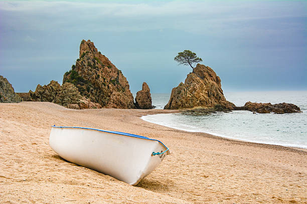 Image showing the tranquility of Mar Menuda beach in Tossa de Mar at dawn on a cloudy day, with a white boat and a rock formation with a solitary tree in the background.
