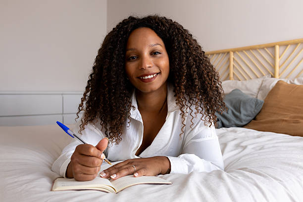 istock Young woman lying on bed writing in notebook, smiling at camera 2214843586
