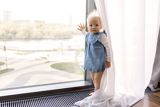 Curious little child standing near large window looking outside. Cute infant exploring visible world and nature, wanting to go outdoors, touching glass