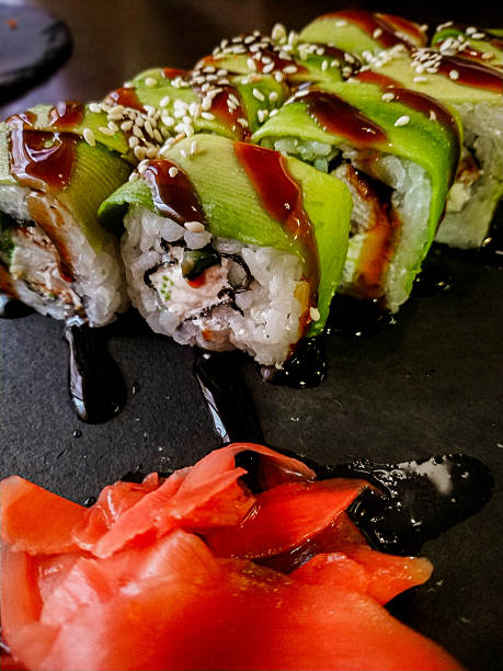 istock Rolls with eel and philadelphia cheese wrapped with thinly sliced ​​avocado and topped with teriyaki sauce on a black stone plate, orange pickled ginger in the foreground, asian food concept 2214858158