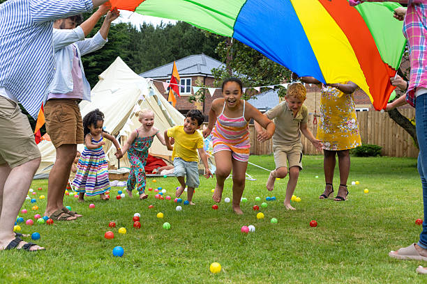 A wide front view of a cheerful group of young children and parents playing together outdoors, at a birthday party on a sunny day. They are enjoying a fun summer birthday garden party together with family and friends. They are playing with a parachute and plastic balls, the young children are running underneath the colourful parachute.\n\nVideos are available similar to this scenario.
