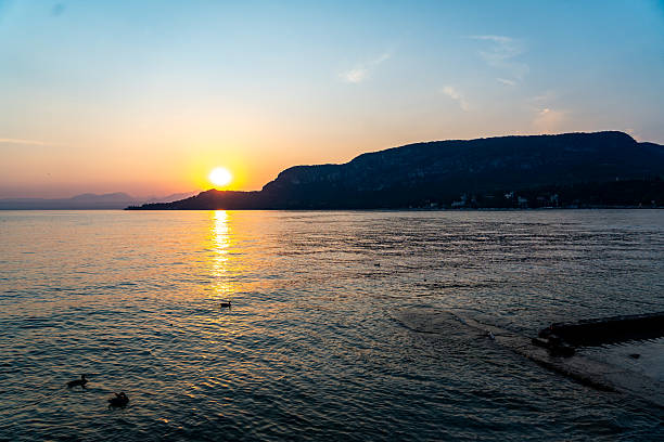 A breathtaking view of the sun setting behind the silhouette of hills and cypress trees along the shore of Lago di Garda, Italy. The sky is bathed in warm hues of orange and yellow, with the golden sunlight reflecting across the calm lake surface, creating a peaceful and romantic atmosphere.