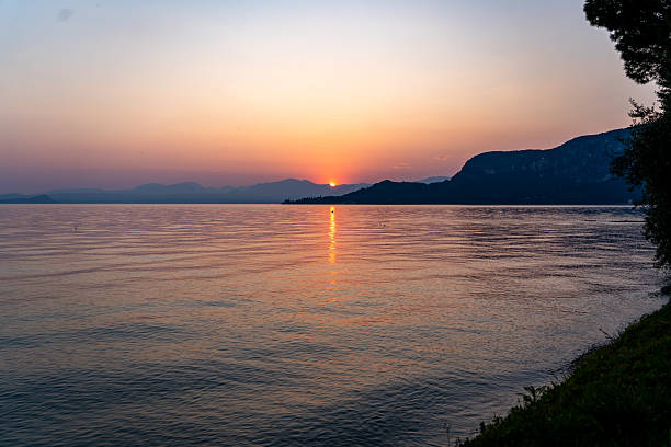 A breathtaking view of the sun setting behind the silhouette of hills and cypress trees along the shore of Lago di Garda, Italy. The sky is bathed in warm hues of orange and yellow, with the golden sunlight reflecting across the calm lake surface, creating a peaceful and romantic atmosphere.