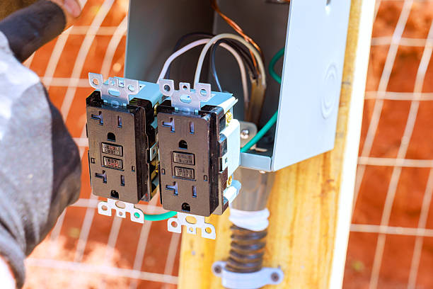 Electrician installing dual electrical outlets in circuit box located outdoors on wooden post during daylight
